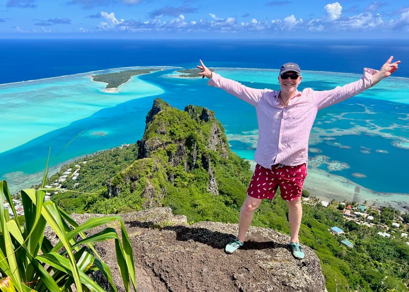 Alan Peterson, arms raised, on a rocky summit high above Maupiti's lagoon and reef.