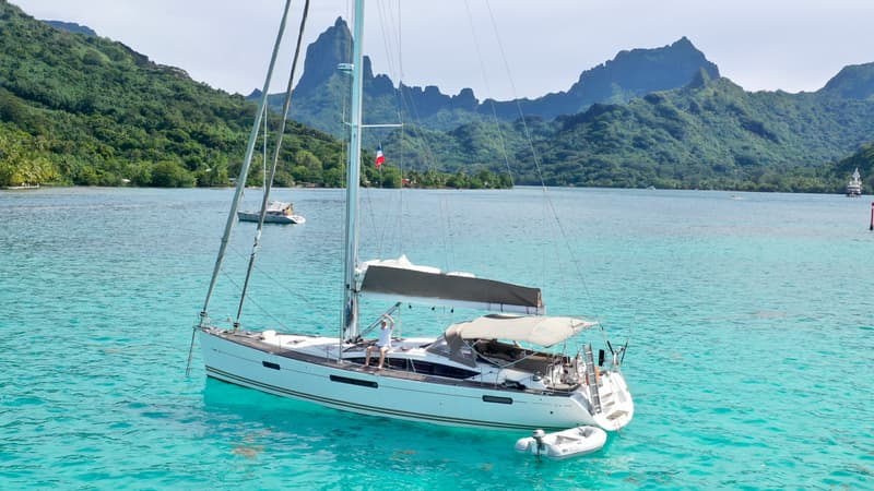 Aerial view of Cyrolia at anchor in clear turquoise water, her dinghy alongside, with green mountains behind.