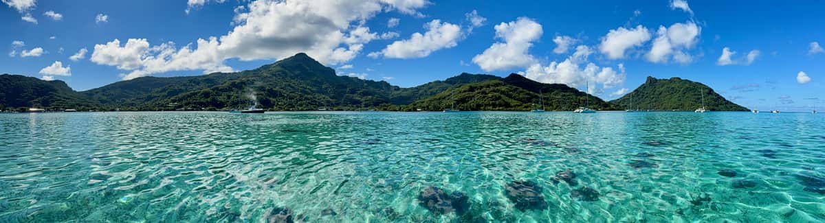 A wide panorama of a clear, shallow lagoon in Huahine, with green hills and anchored sailboats along the far shore.